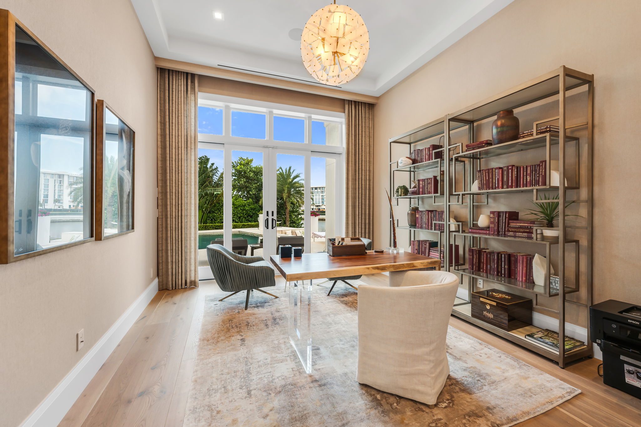 Modern home office with wooden desk with acrylic legs, three plush chairs, large bookshelf with books and decor, soft rug, and large windows opening to a pool and garden.