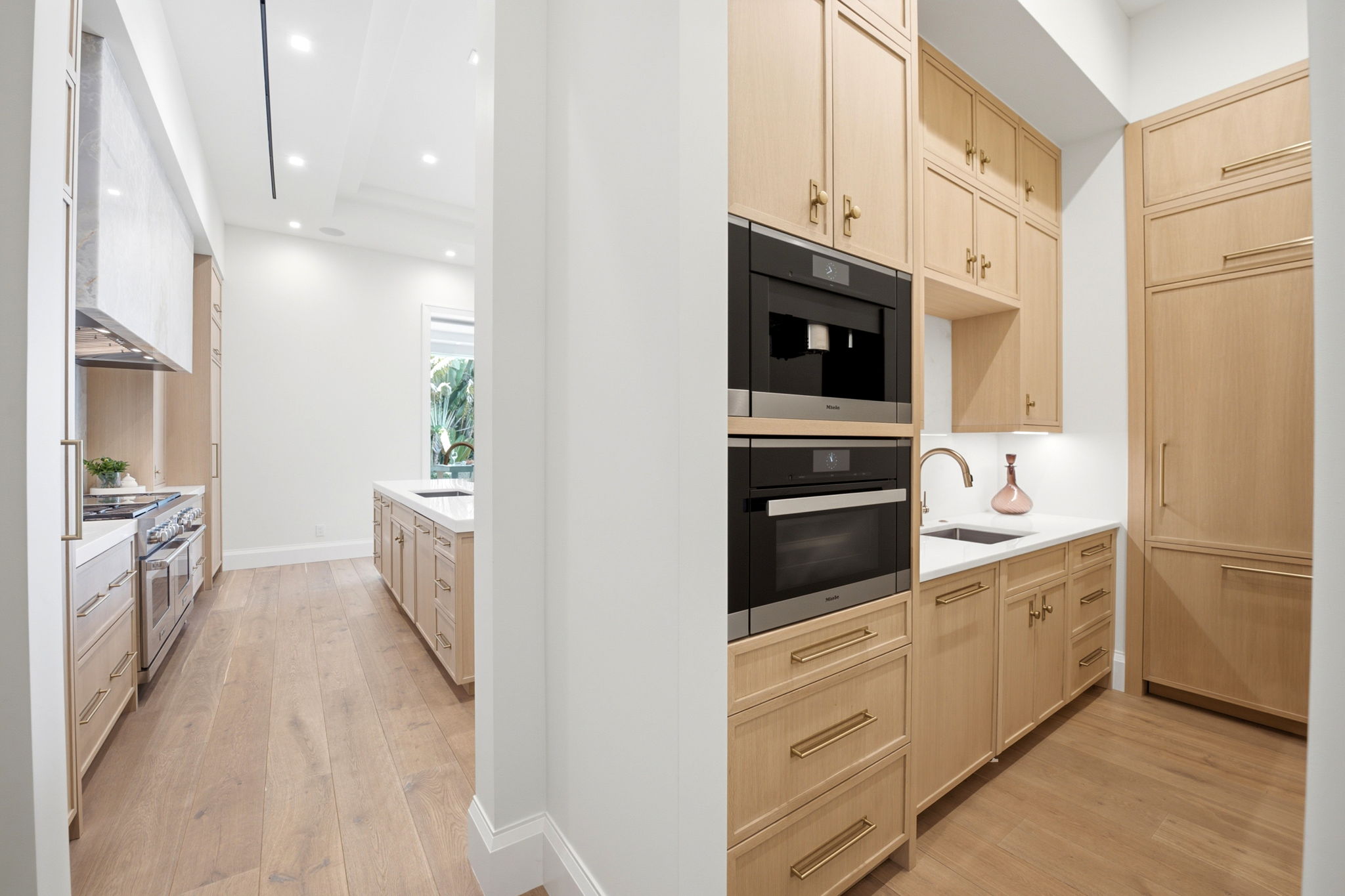 Modern kitchen with light wood cabinets, stainless steel double oven, built-in refrigerator, white countertops, under-cabinet lighting, and wide plank wood flooring.