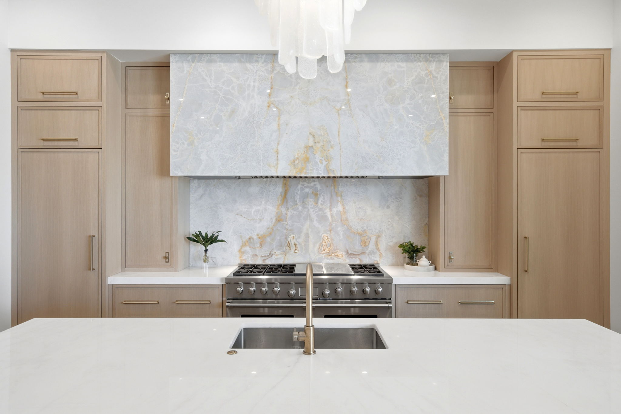 Modern kitchen with light wood cabinets, a white marble countertop island with an integrated stainless steel sink and brass faucet, and a large stainless steel stove beneath a marble backsplash and vent hood featuring white and gold veining, accented by small green plants on either side of the stove.