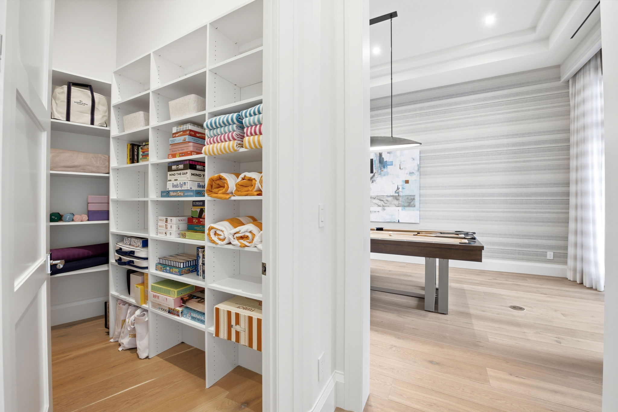 Open door reveals organized storage room with white shelves holding board games, rolled towels, and storage bins, next to a game room with a pool table, abstract wall art, and striped wallpaper.