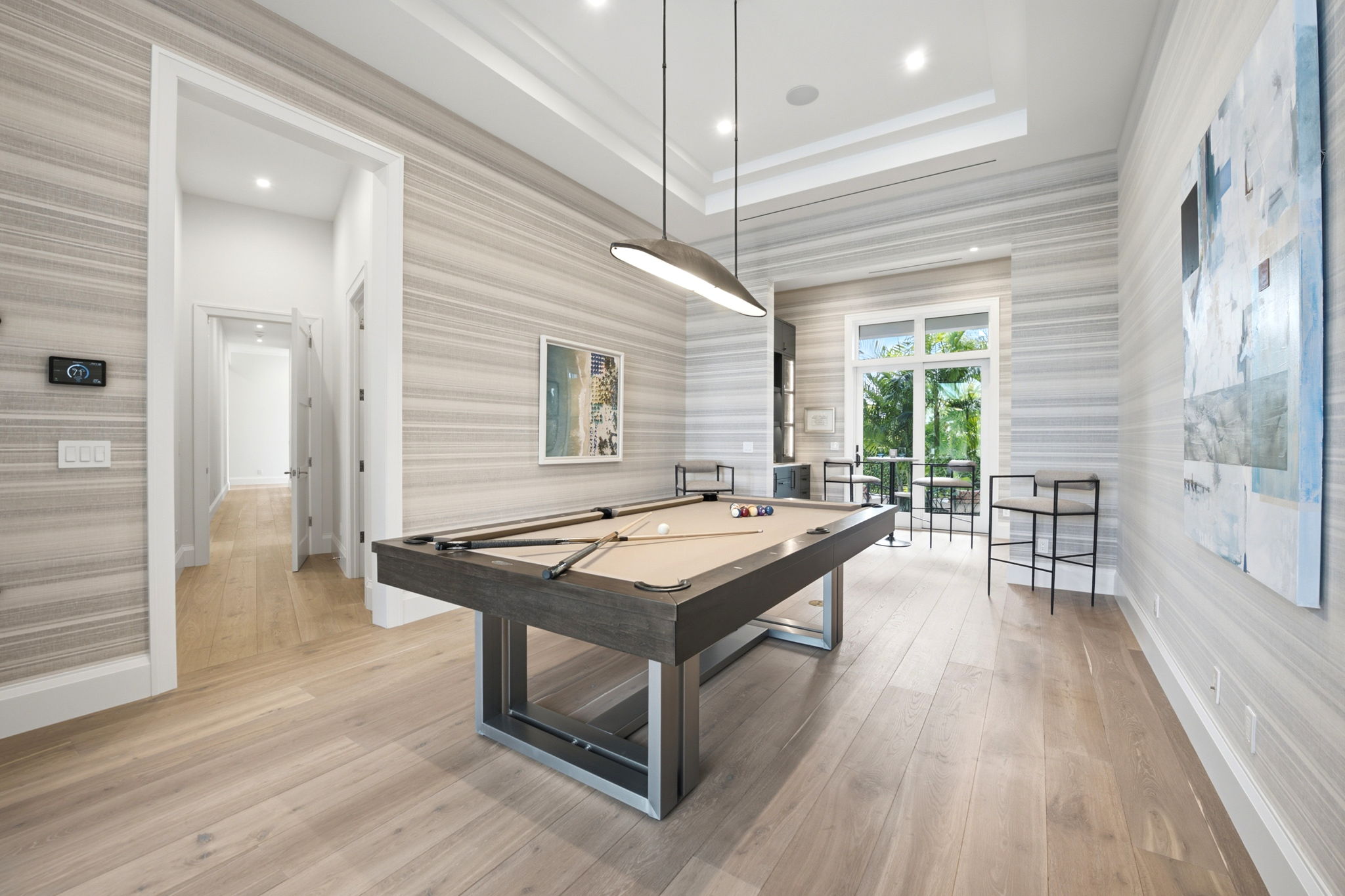 Bright modern room with wooden floor and striped gray wallpaper featuring a pool table with cues and balls, two high chairs, a small bar table near glass doors opening to outdoor greenery, artwork on walls, and a unique ceiling light above the table.