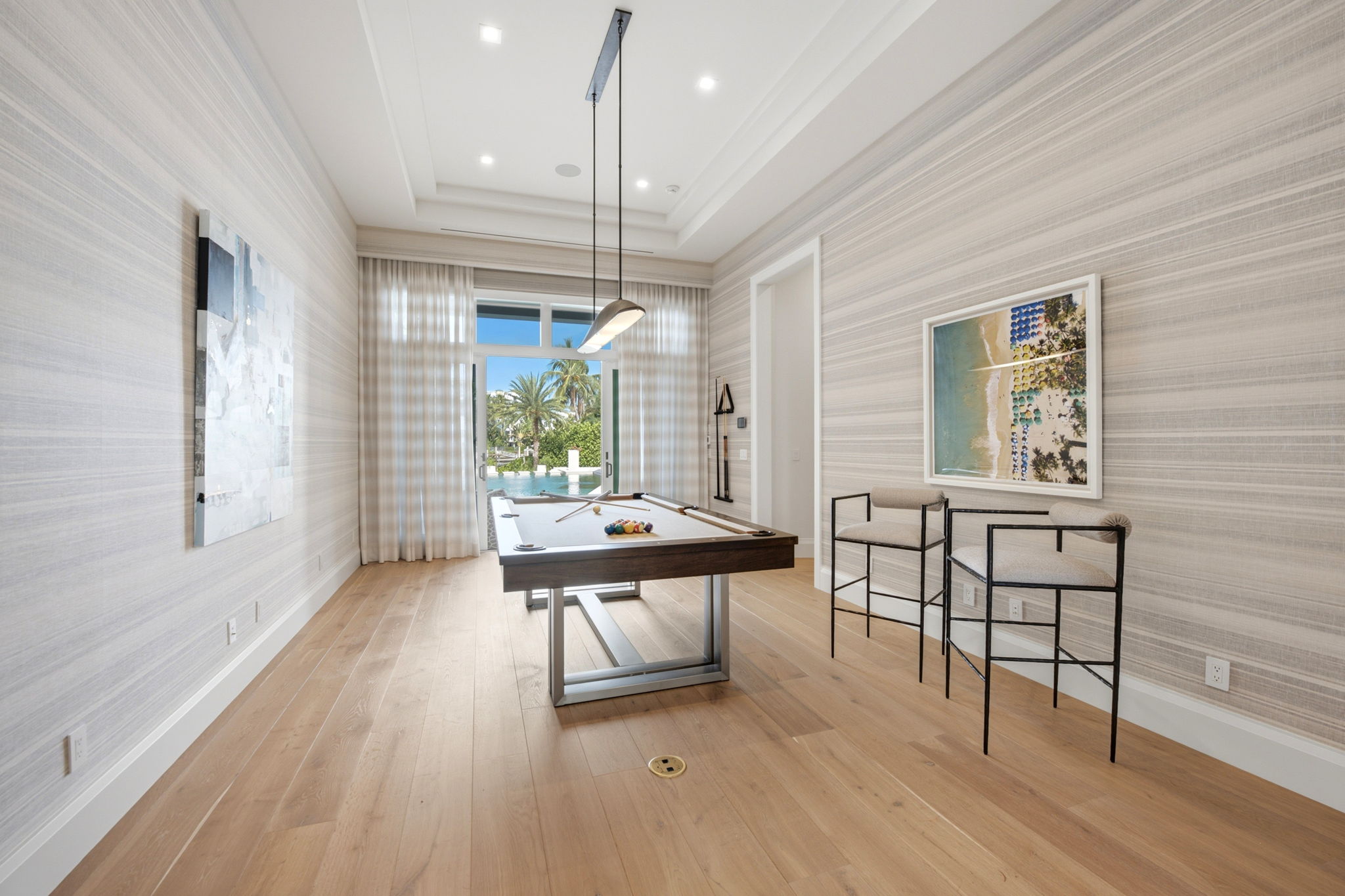 Bright game room featuring a modern pool table with a cue stick and billiard balls, two stylish bar stools with metal frames and light cushions, beach-themed artwork on walls, and large sliding glass doors opening to a pool and palm trees outside.