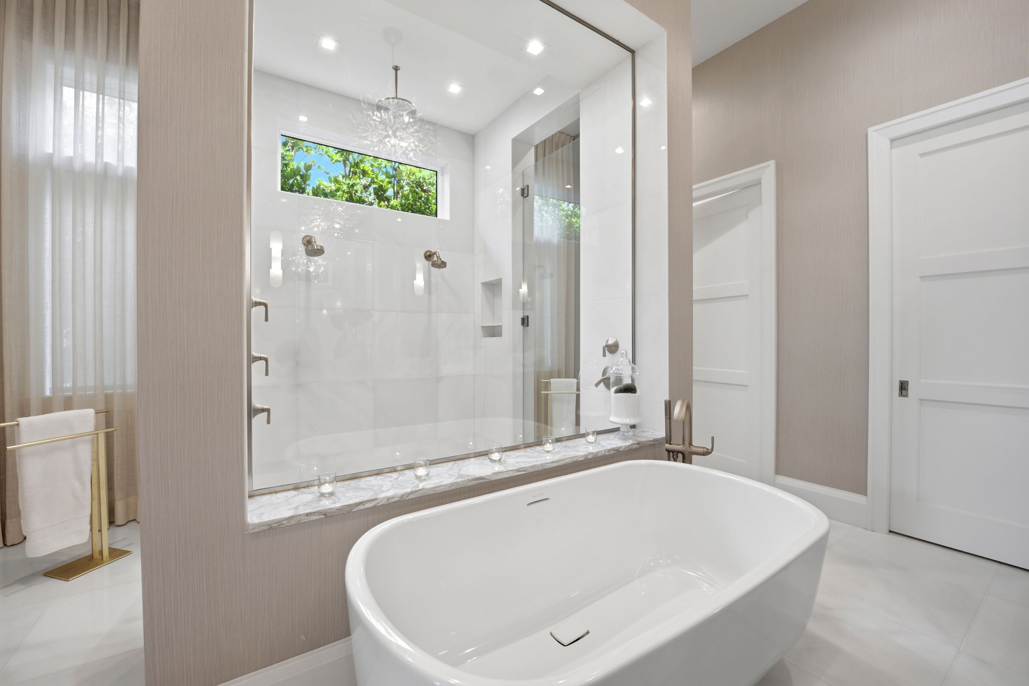 Modern bathroom featuring a white freestanding bathtub in front of a large framed glass partition that reveals a shower area with double showerheads and a high rectangular window showing green foliage outside, with neutral wall colors, white doors, and a gold towel rack holding a white towel.