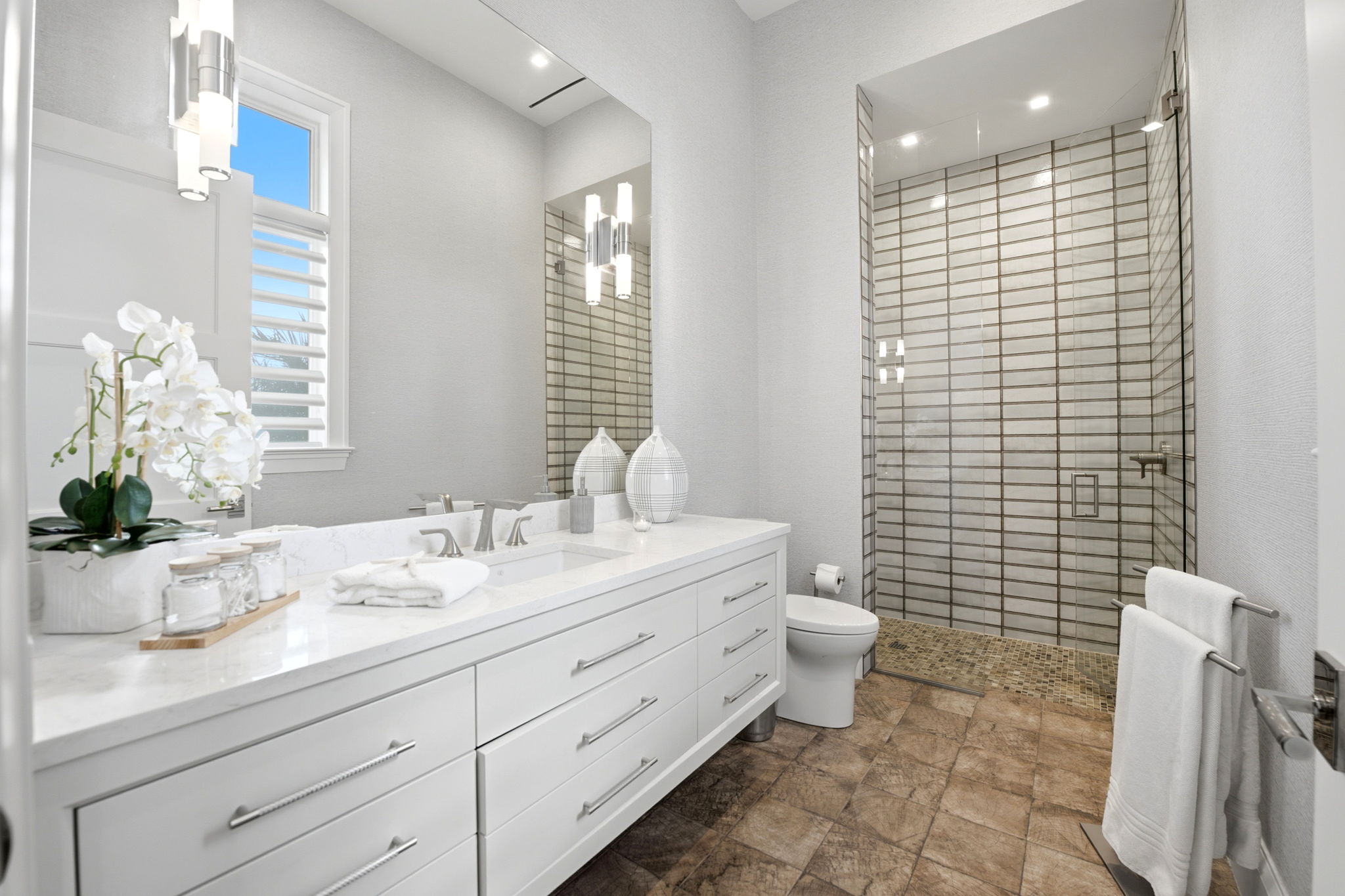 Modern bathroom with white cabinetry and a marble countertop featuring a sink, decorative vases, a soap dispenser, and a small towel, next to a window with shutter blinds. A glass-enclosed shower with tiled walls and mosaic floor is beside a white toilet. Two white towels hang on a chrome towel rack on the right wall. The floor is covered with brown stone tiles.