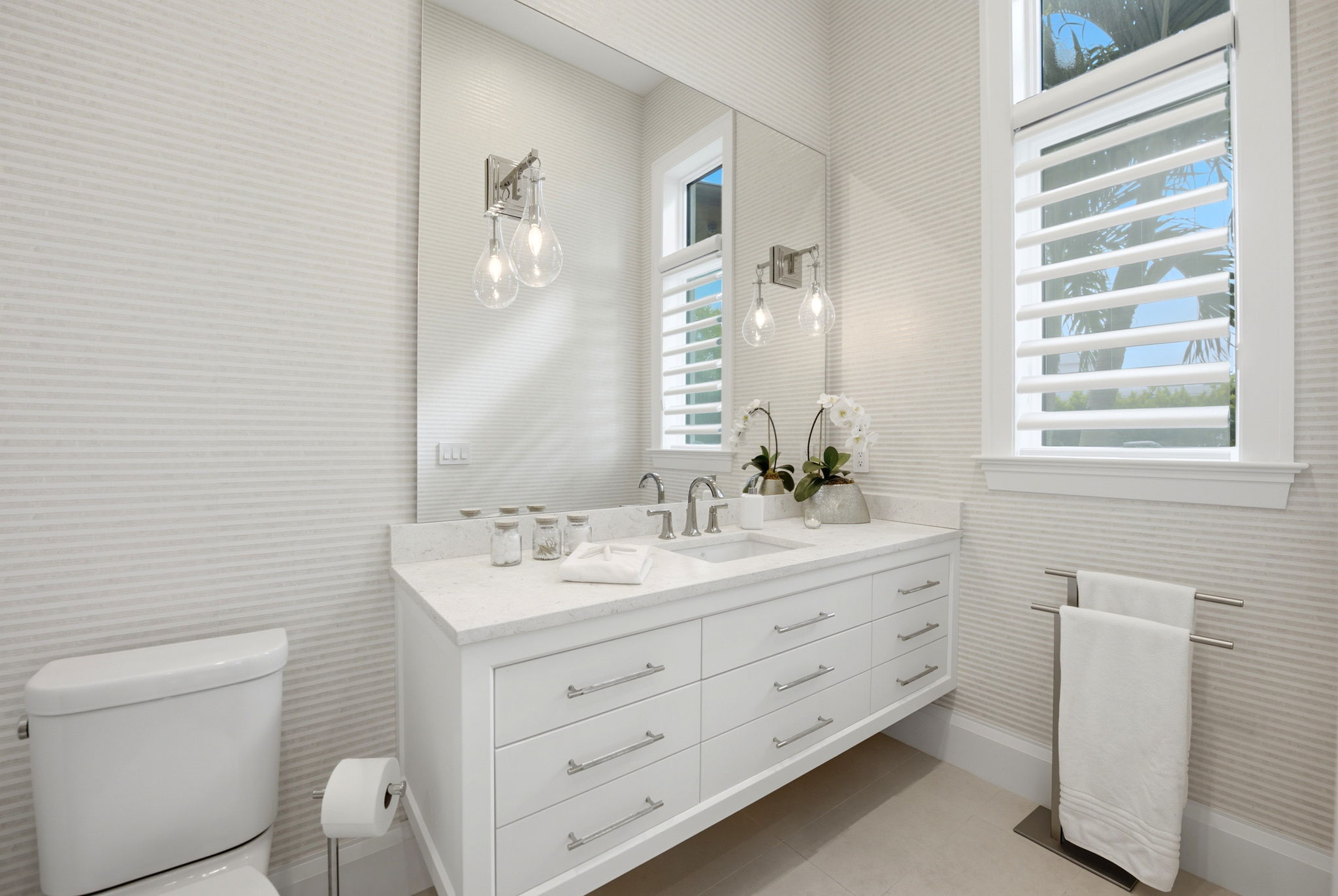 Bright modern bathroom with white vanity featuring multiple drawers, a rectangular sink, large mirror, two hanging bulb light fixtures, a window with white shutters, white toilet with a freestanding toilet paper holder, and a towel rack with white towels.