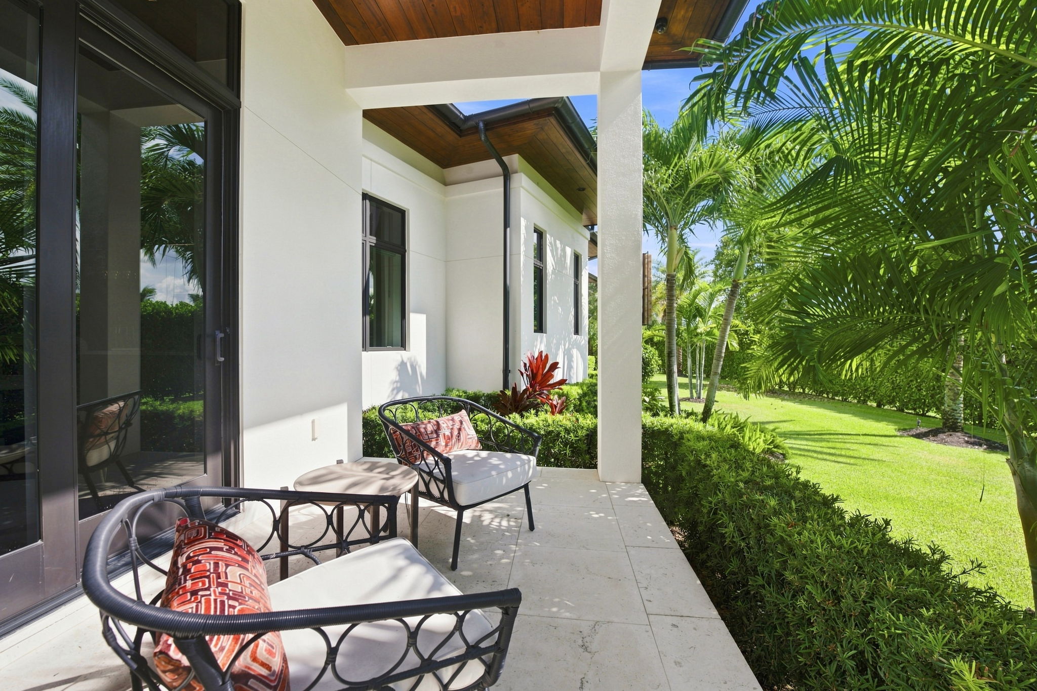 Outdoor patio with two black metal chairs with white cushions and red patterned pillows, a small round table, white tiled floor, white house wall, wooden ceiling, and lush green palm trees and bushes in the background.