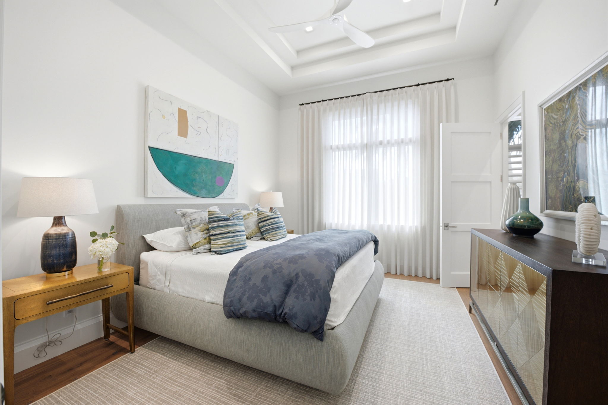 Bright modern bedroom with a light gray upholstered bed topped with white and striped pillows and a blue patterned comforter, beige rug, wooden nightstand with lamp and flowers, large window with sheer white curtains, dark wood dresser with decorative vases, and a ceiling fan in a recessed ceiling.