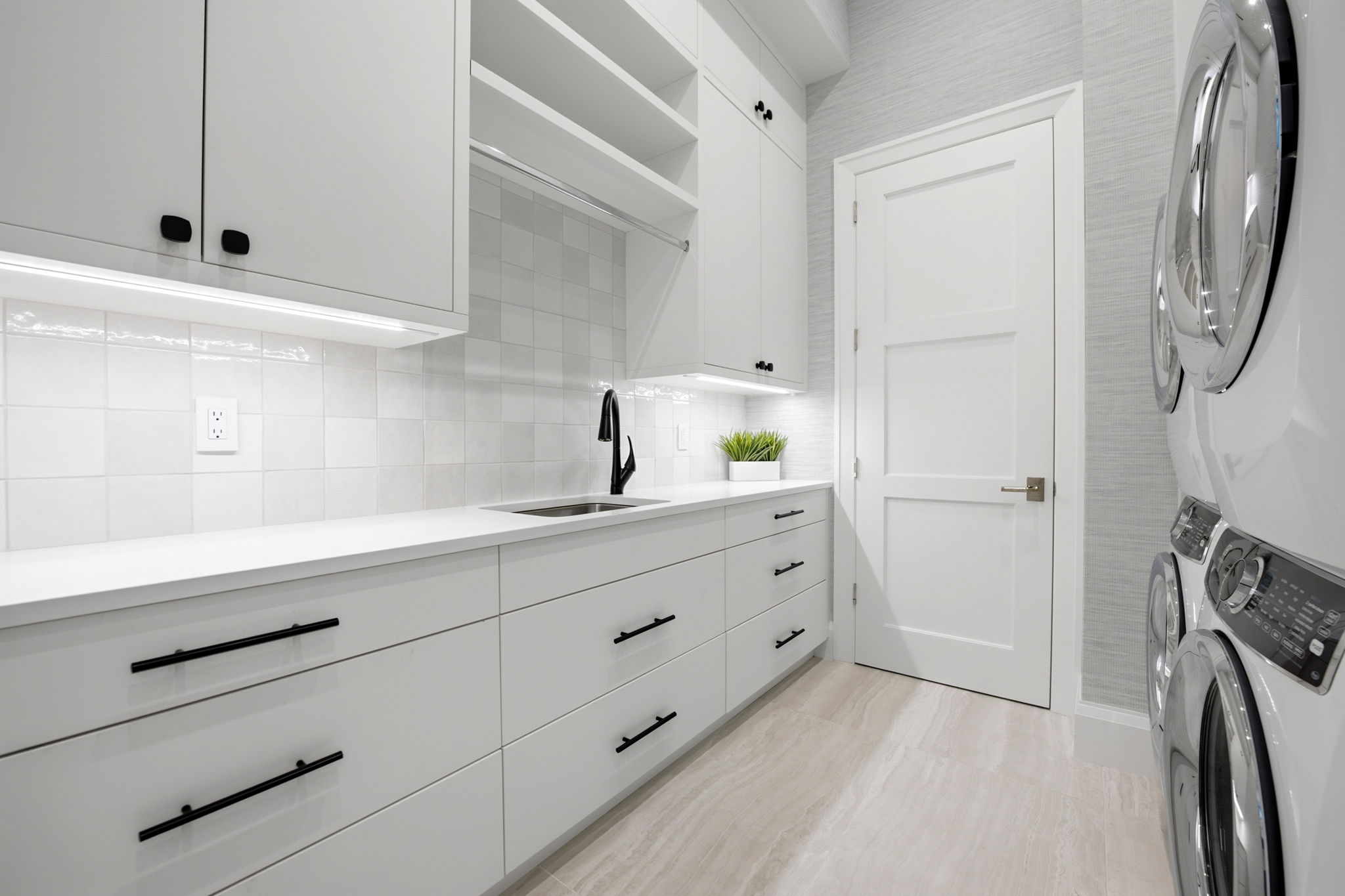 Modern laundry room with white cabinetry featuring black handles, a black sink faucet, white tiled backsplash, white countertop with a small green plant, and stacked white washer and dryer units on the right side.