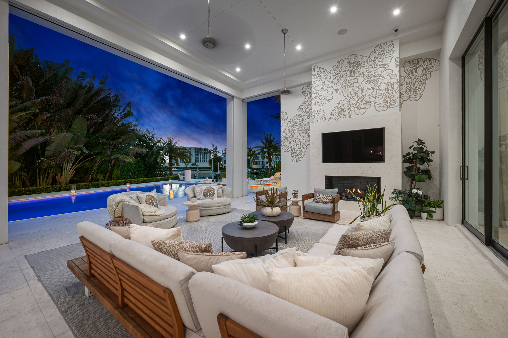 Spacious modern outdoor living area at night with a large beige sectional sofa, round coffee tables, two beige swivel chairs, a gray armchair, fireplace with TV above, potted plants, and a view of a swimming pool and tropical plants under a deep blue sky.