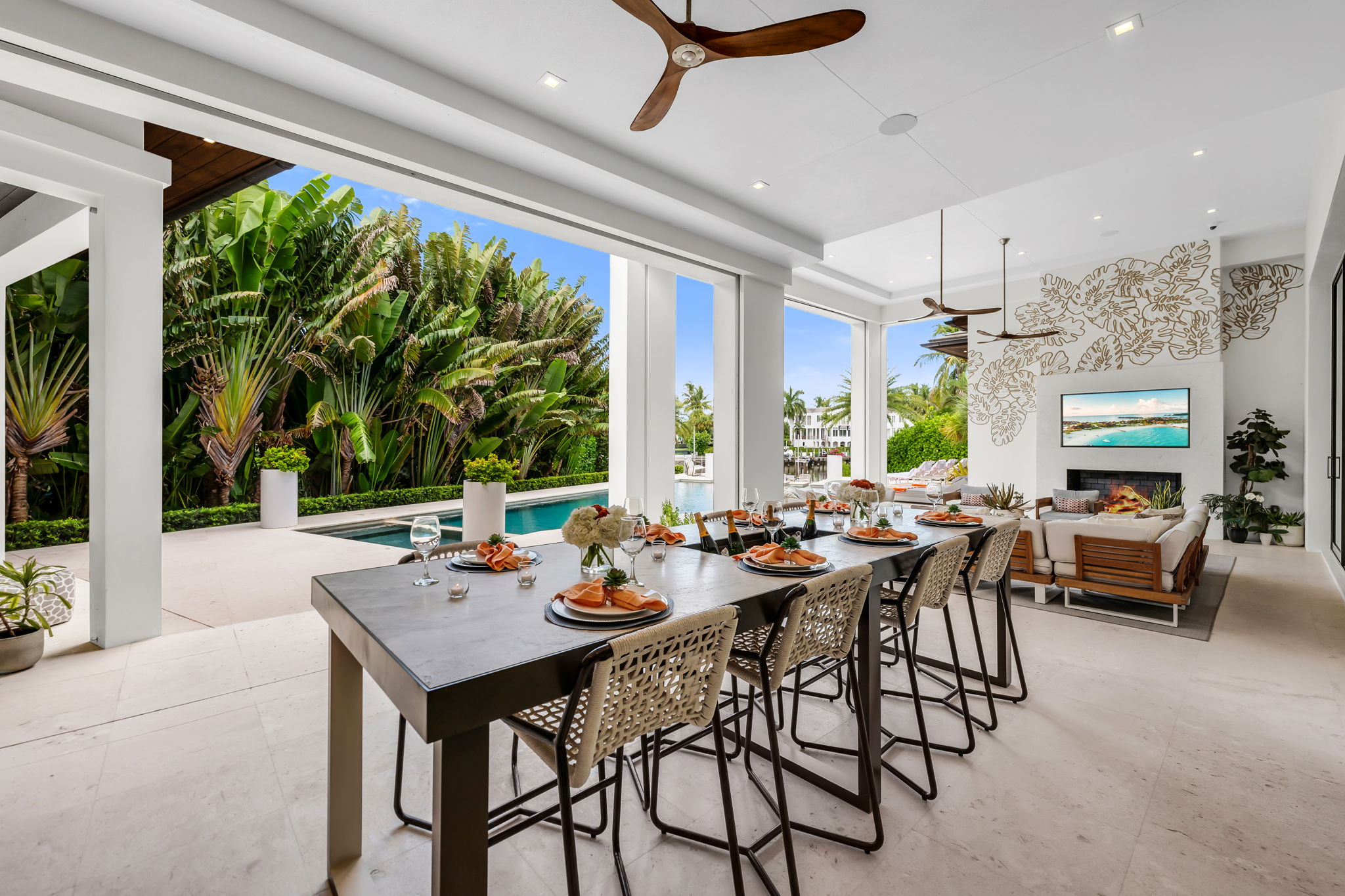 Covered outdoor dining and lounge area with a long modern dining table set with plates, napkins, and wine glasses, a ceiling fan, decorative wall with a TV above a fireplace, and a pool surrounded by lush greenery.