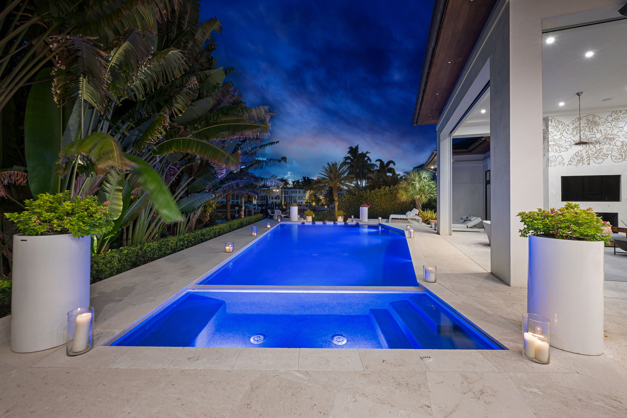 Modern outdoor swimming pool illuminated with blue lights at night, surrounded by tropical plants and large white planters with greenery, with a covered patio area and lounge chairs visible on the right.