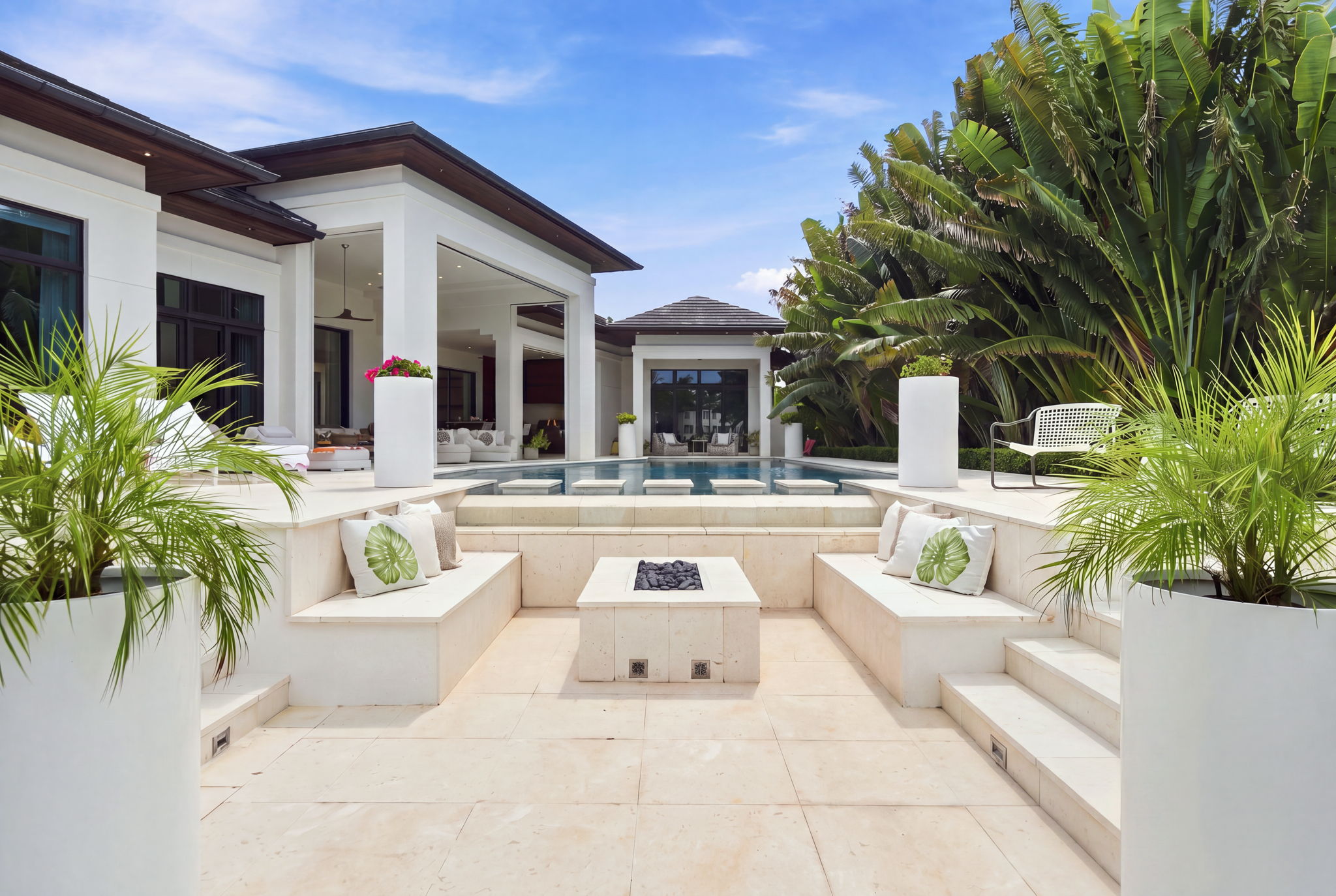 Modern outdoor patio with built-in beige stone seating around a rectangular fire pit, decorative cushions with green leaf prints, large planters with palm plants, a rectangular pool with stepping stones, and a white house with large windows and lush tropical greenery in the background under a partly cloudy sky.