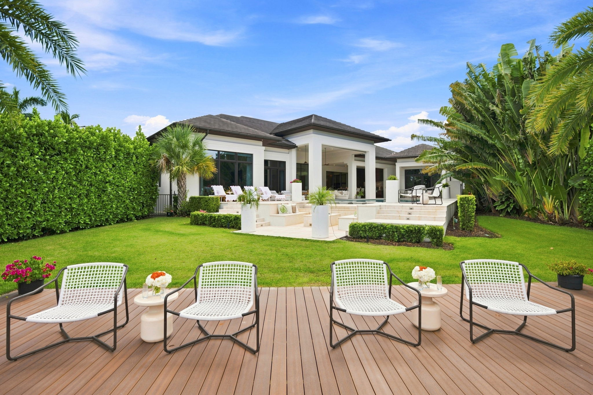 Modern white house with a large manicured green lawn and tropical plants, featuring a wooden deck in the foreground with four white woven chairs and two small side tables with floral arrangements.