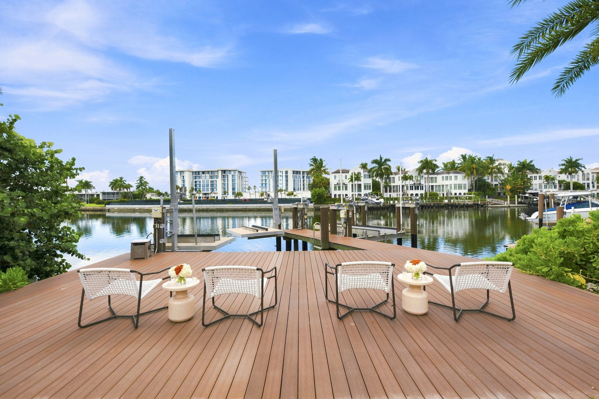 Wooden dock with four white woven chairs and two small round tables with flower arrangements, overlooking calm water with boat lifts, palm trees, and white buildings under a blue sky.