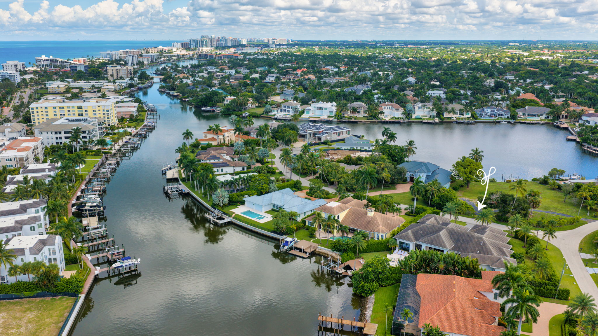 Aerial view of a waterfront residential neighborhood with houses, palm trees, docks, calm canal water, and a lake extending into the distance under a partly cloudy sky.