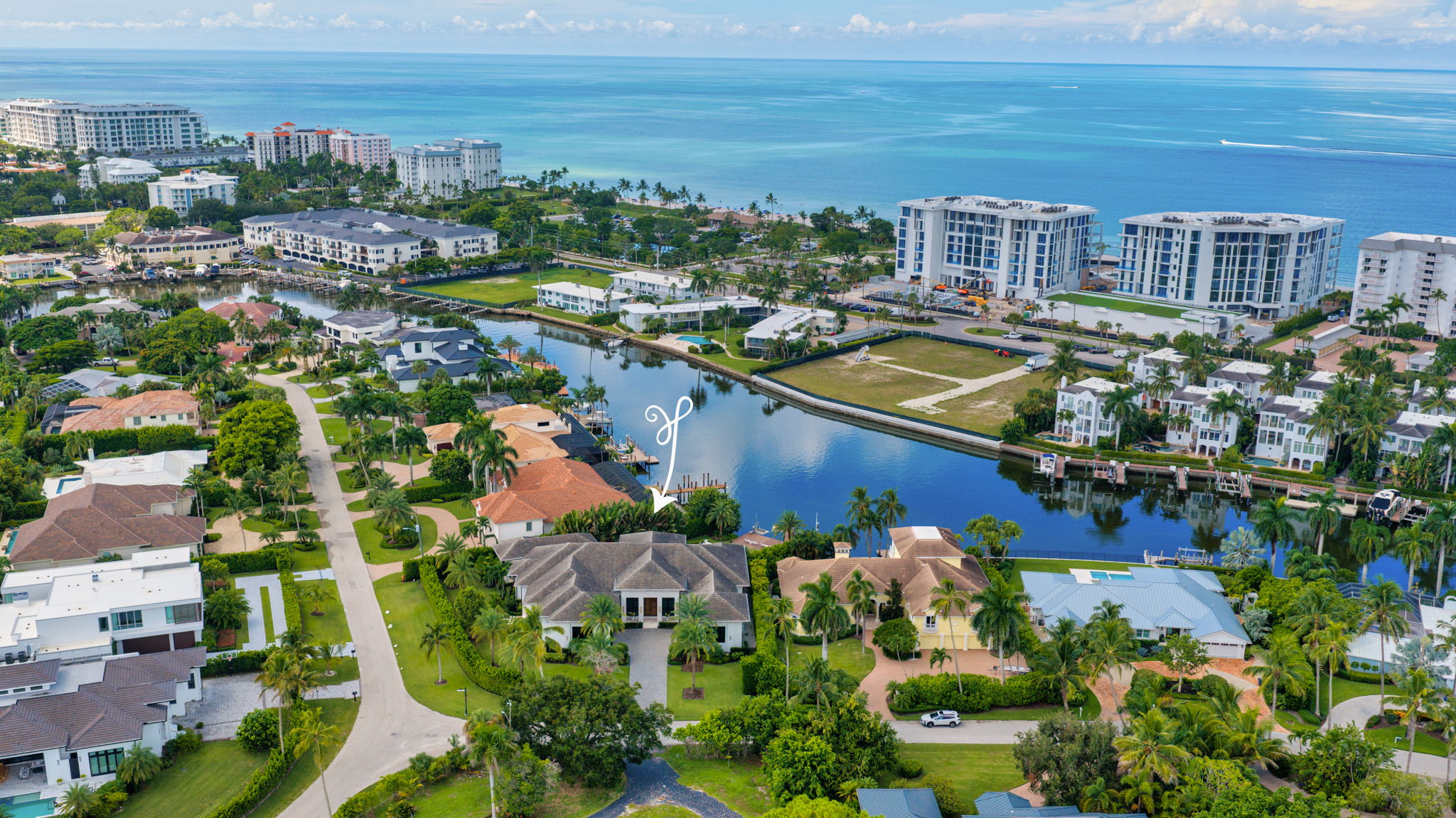 Aerial view of a coastal residential neighborhood with mansions, palm trees, a canal, and the ocean in the background under a partly cloudy sky.