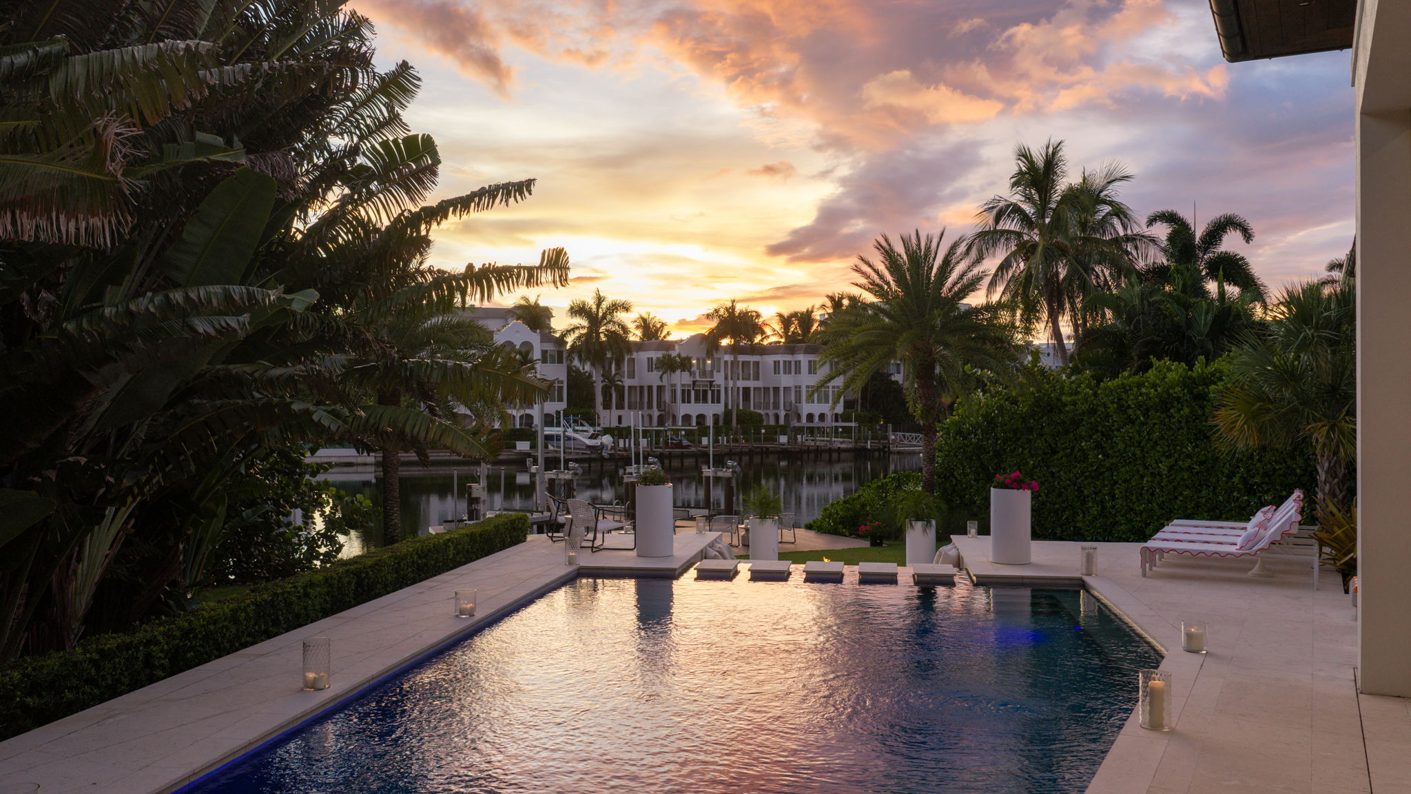 Outdoor swimming pool at sunset with candles placed around its edge, surrounded by tropical plants and palm trees, with a canal and white residential buildings in the background.