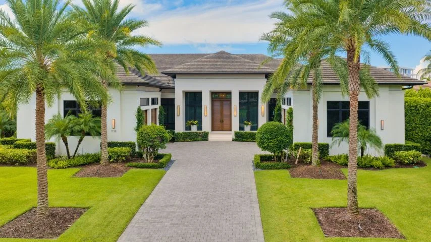 Modern single-story house with white walls, a gray shingled roof, and a central wooden door flanked by large windows, surrounded by landscaping with palm trees and neatly trimmed bushes along a paved driveway.