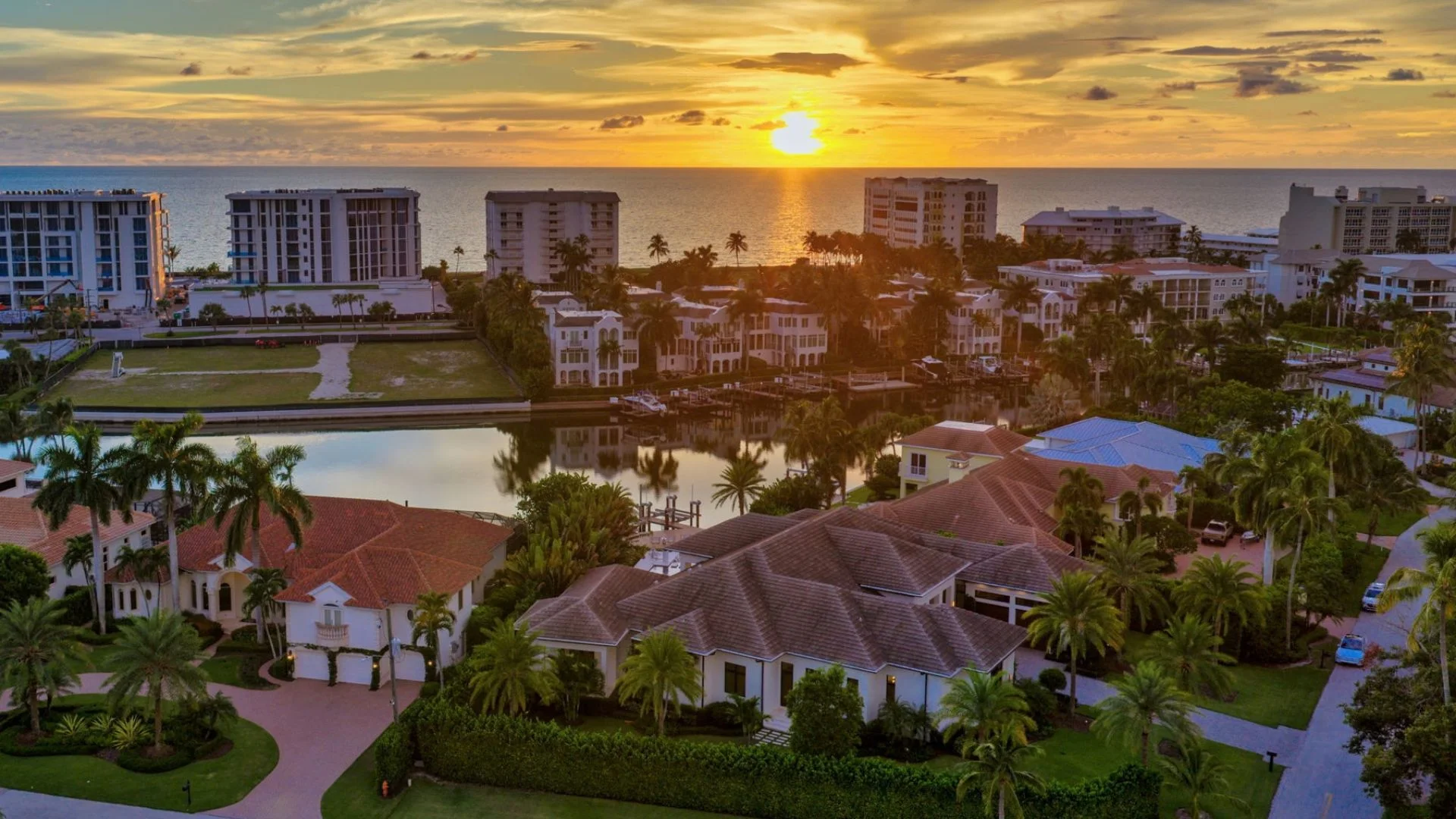 Aerial view of a coastal residential area at sunset with large houses featuring tiled roofs, palm trees, a canal with boats, and high-rise buildings along the ocean shoreline reflecting the golden sunlight.