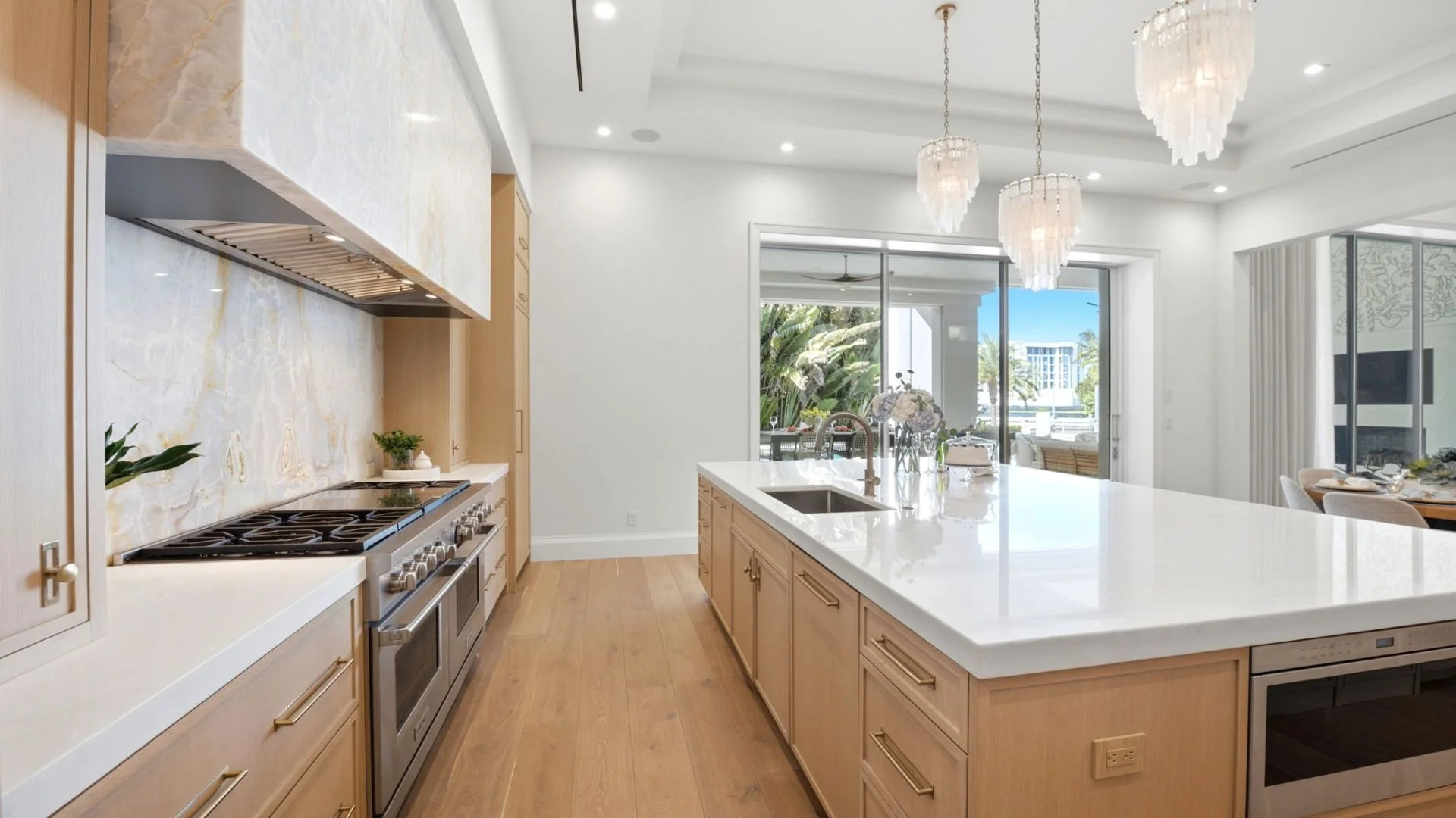 Modern kitchen with light wooden cabinets, white marble countertops, large island with sink, stainless steel stove, glass-pendant lights, and sliding glass doors leading outside.
